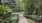 Concrete path at Marq at the Pinehills winding through a green garden with trees, shrubs, grassy area, and outdoor seating.