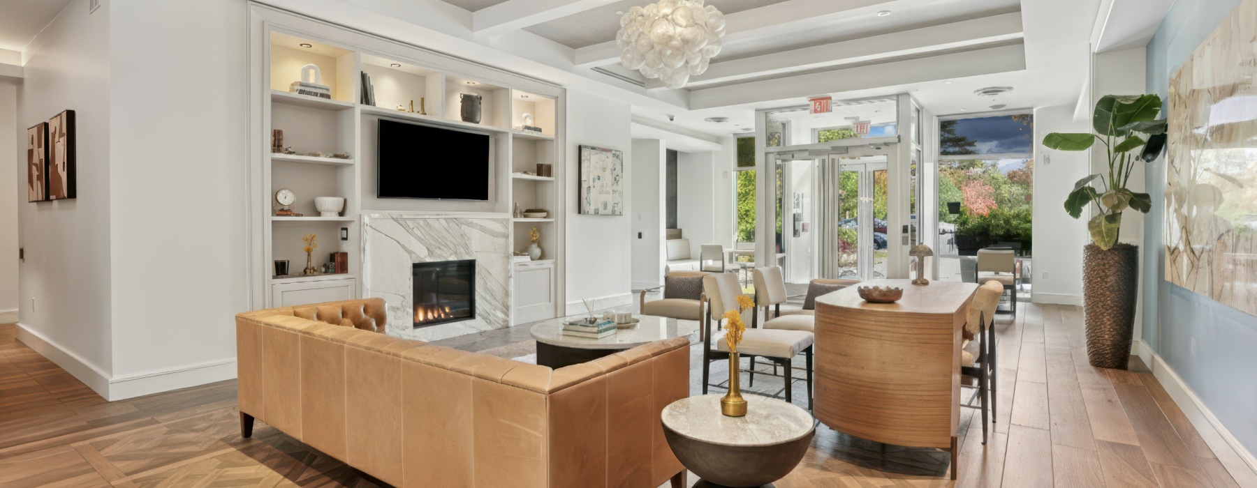 Lounge area at Marq at the Pinehills with fireplace, wall-mounted TV, built-in bookshelves, white sofa and plants.